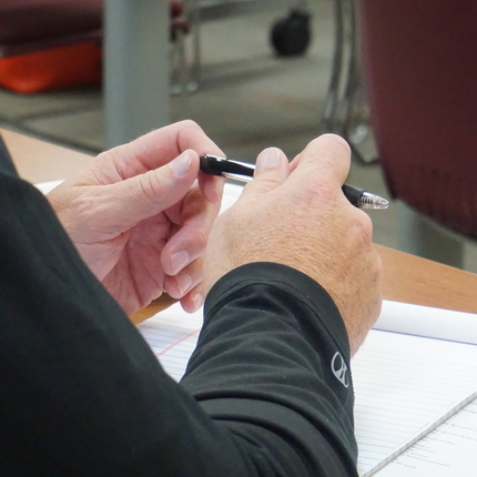 A person's hands with black long sleeves holds a black pen with both hands and a white note pad sits on a brown table below