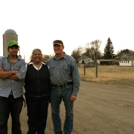 Three individuals - two men and a woman, standing on a farm in front of a grain bin with a house to the right