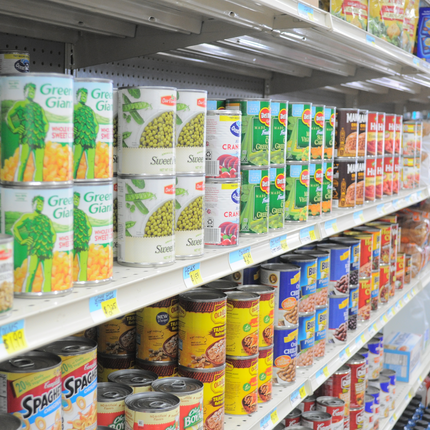 A close-up view of canned goods in a grocery store aisle. We see canned corn, sweet peas, cranberry sauce, Manwich sloppy joe mix, and Spaghetti O's.