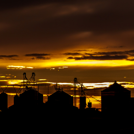 La silueta de tres grandes silos de grano, dos silos de grano más pequeños y dos graneros o dependencias se recorta contra una puesta de sol con intensos tonos naranjas y amarillos, entre nubes oscuras.