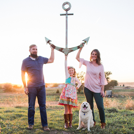 Family portrait of a man, woman, daughter, and dog. All are touching an anchor in a pasture.