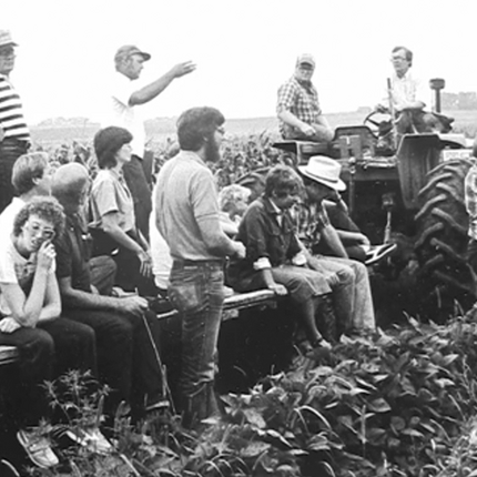 Fotografía histórica en blanco y negro de un grupo de agricultores y miembros de la comunidad reunidos en un campo de cultivo, sentados y de pie sobre equipos agrícolas y un tractor, mientras un hombre hace un gesto como si hablara a la multitud.