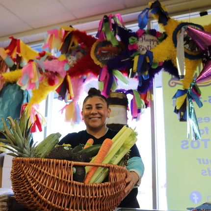 Woman stands behind counter with basket of vegetables in front of her and pinatas behind her