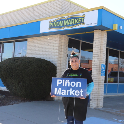 Woman stands with sign in front of a building that says Pinon Market