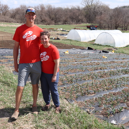 Man and woman in red t-shirts standing in a planted field with white tent greenhouses in background.