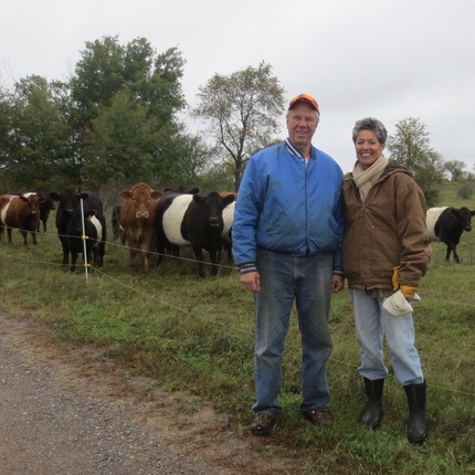 A man and a woman stand in a field smiling with cattle behind them