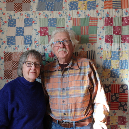 A man and a woman standing in front of a quilt in a 1800s farm house