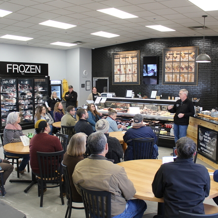 A group of people sitting at tables in a restaurant, with a man talking at the front of the room. The man is standing in front of cases holding meat for sale.