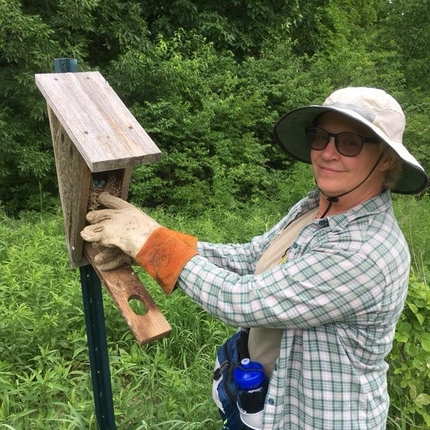 Woman in garden hat and heavy gloves, standing in front of a bird house box open containing a nest and eggs