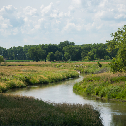 body of water running through a field with landscape and trees on both sides and in the distance.