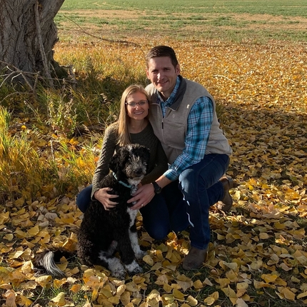 Couple kneeling pose with a black and white dog near a tree with yellow leaves on the ground.