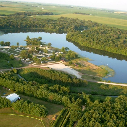An aerial shot of a lake surrounded by green trees, then beyond that, green fields. On the left side of the lake is a campground with white roads and white camper pads, with a handful of travel trailers parked in spots.
