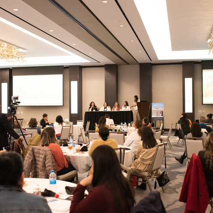 group of people sitting at tables, listening to panel at front