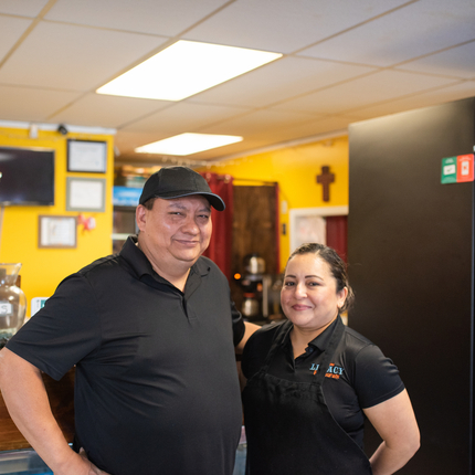 Man and a woman with an apron stand next to a beverage cooler in a restaurant with yellow and orange walls in background