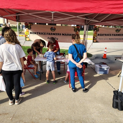 Several adults and several children gathered under a red tent. A sign behind the people reads, "Beatrice Farmers Market"