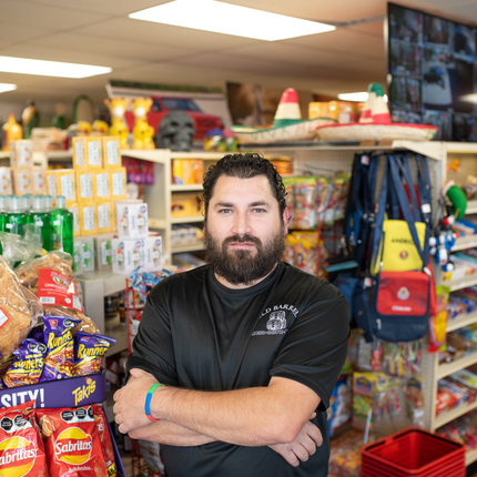 Hombre con barba y camisa negra posa para una foto con brazos cruzados en frente de mercancía en una tienda