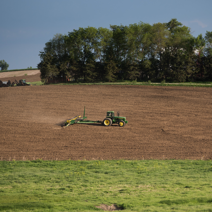 A green John Deere tractor pulls a green planter behind it in a brown, freshly-tilled field. The sky is blue and there is a tree line in the background.