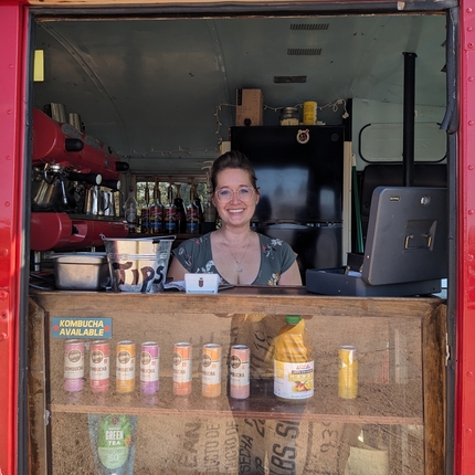 Mujer esta parada detras de un mostrador de bebidas de kombucha y una maquina para hacer cafe expreso a su derecha