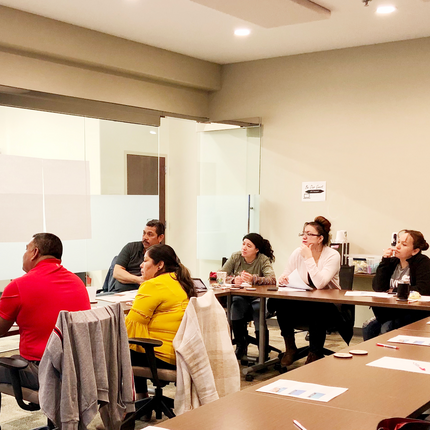 A group of people sit in rolling office chairs around rectangular tables, arranged in a "U" pattern. They appear to be listening to a speaker.