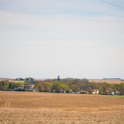 Landscape photo of a harvested field in foreground with small town with fall trees in the middle of the photo. A water tower is in the very center, with its top above the trees.