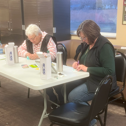 Two women sit at a rectangular table, working on crafts. The woman on the left has short white hair and wears glasses, a pink and white striped hoodie, and a black vest. The woman on the right has shoulder-length brown hair, glasses, and wears a dark green top with a black vest. Both are weeding small pieces of vinyl. On the table are water bottles, stainless steel tumblers, small craft supplies, and white boxes.