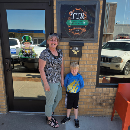 A woman and a young boy stand outside a brick building beneath a sign that reads “TTS Hometown Pub & Grub, Established 2024.” The woman is smiling and wearing a patterned black and white shirt with light green pants, while the boy wears a blue hoodie with a yellow flame design and black shorts. The entrance door next to them has a decal featuring a child in a leprechaun hat and the same business name. 