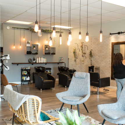 Two grey chairs  along with a wick chair are shown in the middle of a hair salon with light fixtures coming down form the ceiling while a woman in black shirt works on woman's blond hair in the distance