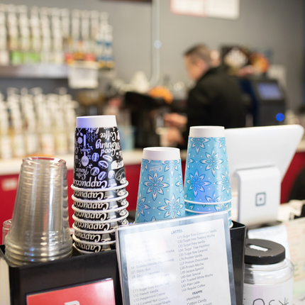 Vasos de distintos colores al frente mientras un hombre al fondo maneja una cafetera।
