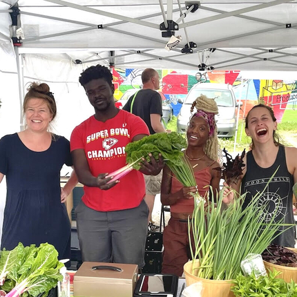 Kelly Alsup and three other people pose for picture smiling with vegetables in hand at farmers market