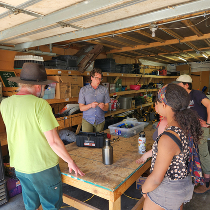 Two white males on the left, a woman with curly hair and tan skin and two other individuals on the right stand around a wooden table with items on it inside an enclosed space with shelving in the background