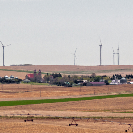 Una vista lejana de un pequeño pueblo en medio de campos marrones cosechados, con turbinas eólicas salpicando las colinas por encima y detrás del pueblo.