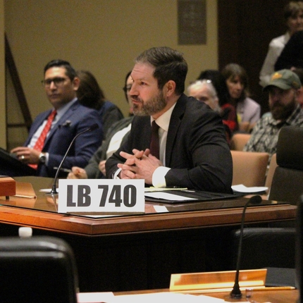 Man in a suit sitting behind wooden desk with a sign on the desk that says "LB 740"
