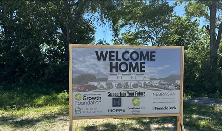 Un cartel temporal que dice "Bienvenido a casa" está en un terreno de tierra con césped verde escaso y árboles frondosos al fondo.