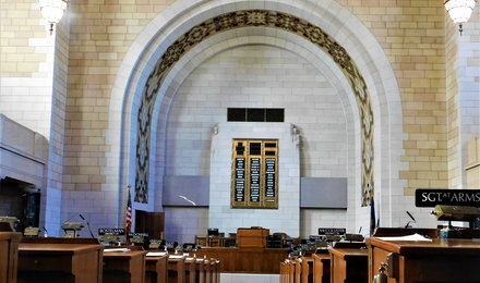 Interior of the Nebraska unicameral legislative room. You are looking up the aisle to the front of the room, wooden benches line each side, and there is an arch at the front of the room with a bench underneath.