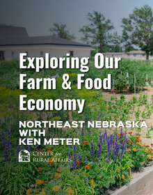 A large community garden with a variety of flowers in a wooden raised bed. In the background, we see a white building. In white letters, it says "Exploring Our Farm & Food Economy Northeast Nebraska with Ken Meter"