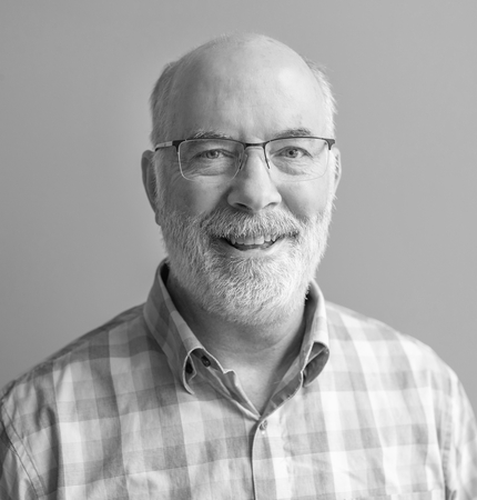 black and white photo of a man, smiling at the camera. Man is bald with short gray hair on either side of his head with a beard and mustache, and wearing glasses. He's wearing a collared checkered shirt.