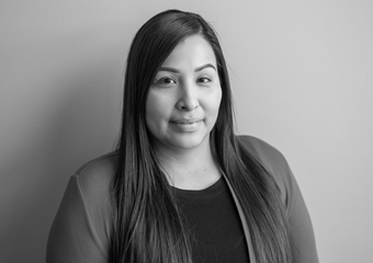Native woman looking directly at the camera with straight dark hair, wearing a black shirt and a lighter sweater over it 