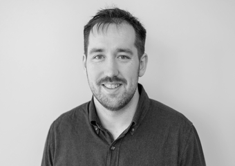 White male with short beard against a grey wall. Black and white photo.