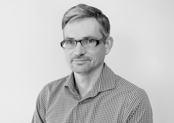 White male with black glasses standing against a white wall. Black and white headshot.