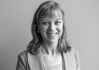 Woman with light colored hair, shoulder length, plus bangs, smiles directly at the camera. She is wearing a plaid jacket, light colored shirt, and pearls.