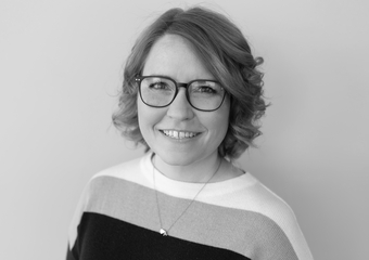 black and white photo of a woman smiling at the camera, with light shoulder-length hair and dark-rimmed glasses