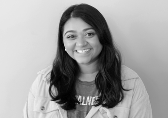 Woman with darker skin smiling at the camera. Has dark wavy hair below her shoulders, and wearing a tan jacket with a darker shirt underneath