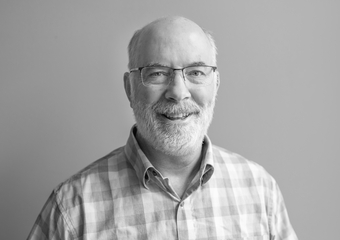 black and white photo of a man, smiling at the camera. Man is bald with short gray hair on either side of his head with a beard and mustache, and wearing glasses. He's wearing a collared checkered shirt.