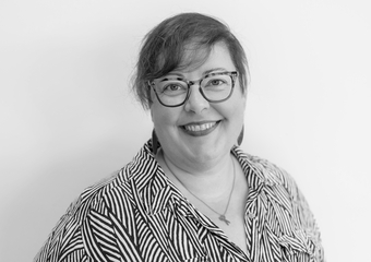 Woman with short hair, smiling at the camera. She is wearing tortoise-shell glasses, has dark lipstick, is wearing a necklace with a pair of scissors charm, and is wearing a leaf imprint top.