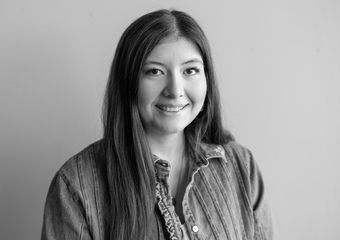 Woman with dark straight hair, looking straight at the camera, smiling. She is wearing a long sleeve button down shirt.