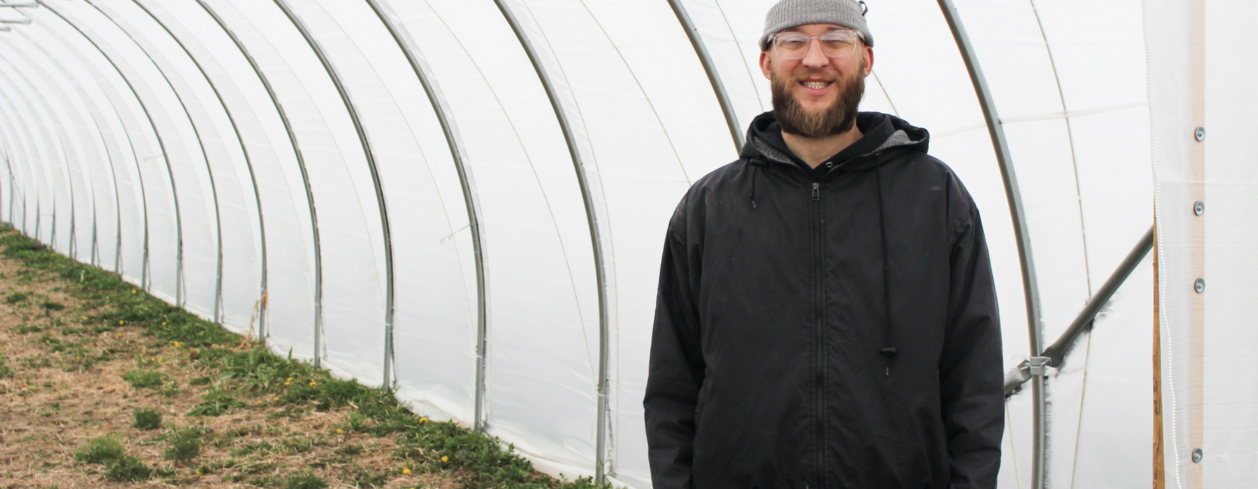 Man in cap and glasses stands smiling in a hoop house.
