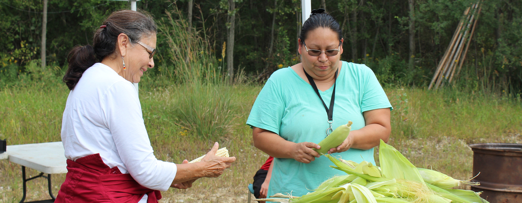Two women shucking Indian corn. Left - a woman wearing a white shirt with a red shirt tied around her waist, with brown hair pulled back, and glasses, smiling. Right: A woman in a blue-green shirt, tied back brown hair, and glasses, frowning at the ear of corn in her hands.