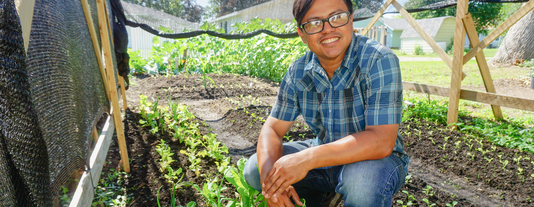 Hispanic male in a blue shirt and jeans squatting in a garden smiling at the camera