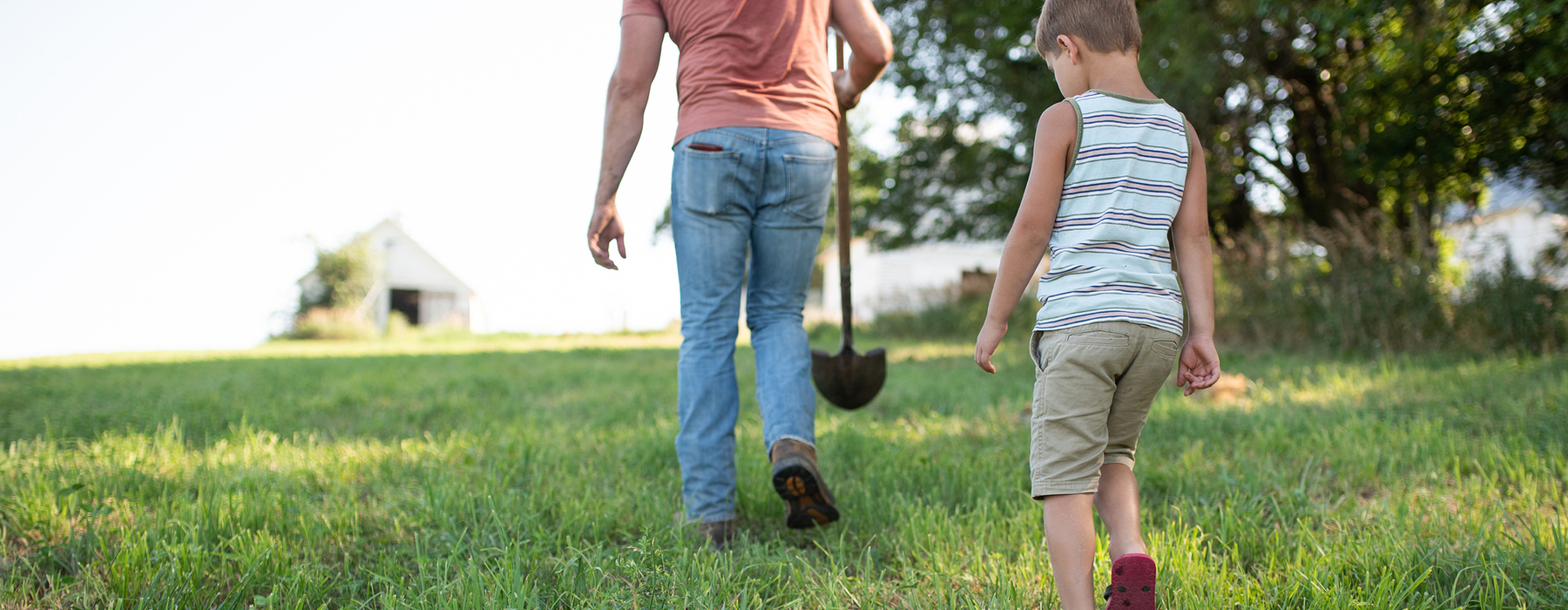 A man in jeans, peach shirt, holding a shovel walks through a grass pasture followed by a young boy in tan shorts and a white and tan striped tank top