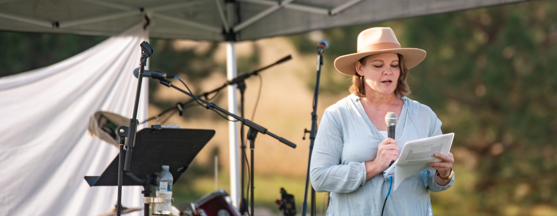 Woman in sunhat and light blue long sleeved shirt speaking into a microphone held in one hand and a couple papers in the other hand
